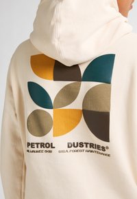 Felpa crema con un design geometrico colorato sulla schiena, caratterizzato da forme blu, verdi, marroni, arancioni e gialle. Sotto, la scritta "PETROL INDUSTRIES".