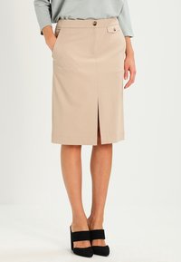 Femme portant une jupe crayon beige arrivant au genou avec fente avant, détail de boutons et chaussures à talons hauts noires.