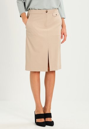 Donna che indossa una gonna a tubino beige di lunghezza al ginocchio con spacco anteriore, dettagli a bottoni e scarpe con tacchi alti nere.