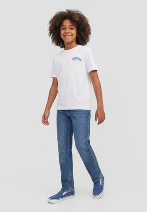 Mosolygó, göndör hajú gyerek fehér pólóban, kék farmerral és kék slip-on cipőben, egy egyszerű háttér előtt áll.