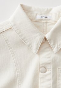 Chemise en denim couleur crème avec un col pointe, des détails de couture visibles et une fermeture à boutons blancs ronds. L'étiquette indique "OPUS."