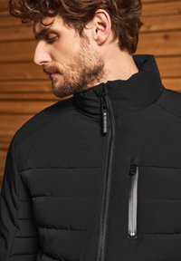 Černá zateplená bunda s vysokým límcem, zipem vpředu a malou náprsní kapsou. Má matný povrch a jemné detaily šití.