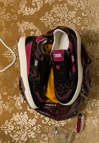 Paire de baskets imprimé léopard et bordeaux sur un tissu doré à motifs, à côté d'un collier de perles, d'un flacon de parfum et d'un étui à rouge à lèvres rouge.
