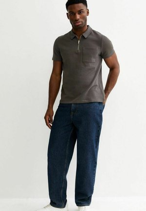 Uomo con polo grigia a maniche corte con taschino sul davanti e jeans blu scuro, in piedi con una mano in tasca contro uno sfondo bianco.