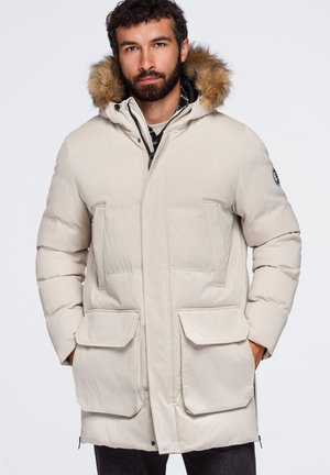 Homme portant un manteau d'hiver matelassé beige avec une capuche bordée de fourrure et de grandes poches avant, les mains dans les poches, debout devant un fond blanc.