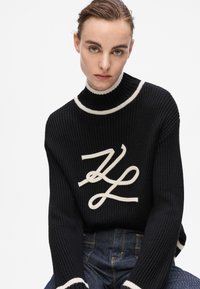 Schwarzer gerippter Pullover mit hohem Kragen, versehen mit einem weißen Logodesign und kontrastierenden weißen Akzenten am Kragen und an den Bündchen.