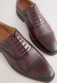 Chaussures en cuir bordeaux avec une tige lisse, des perforations décoratives et un bout arrondi. Des accents marron et un talon légèrement surélevé complètent le design.