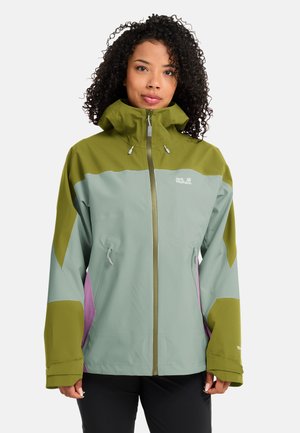 Frau trägt eine grün-hellblaue Outdoor-Jacke mit Reißverschluss, hohem Kragen und Kapuze, steht vor einem einfarbigen weißen Hintergrund.