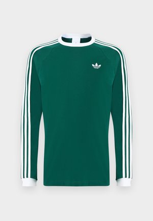 Gari garām piedurknēm, zaļš kokvilnas krekls ar baltu apkakli un aprocēm, ar trim baltiem svītrām uz katras piedurknes un mazu baltu logotipu uz krūtīm.