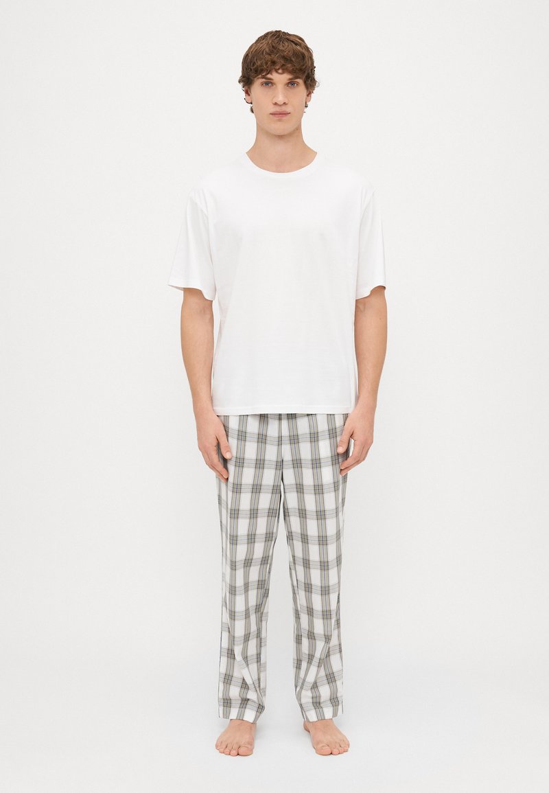 Vit bomull t-shirt med korta ärmar, i kombination med grå rutiga pyjamasbyxor med blå och bruna detaljer. Modellen står barfota.
