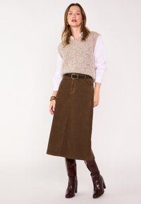 Gilet lavorato a maglia con motivo screziato in beige e marrone sopra una camicia bianca, abbinato a una gonna in velluto a coste marrone e stivali bordeaux alti fino al ginocchio.