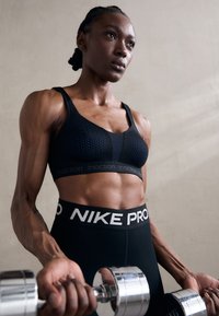 Svart sport-bh med teksturert design som har blå detaljer, sammen med høyt midje svarte leggings med hvit "NIKE PRO" tekst. Sølv hantel.