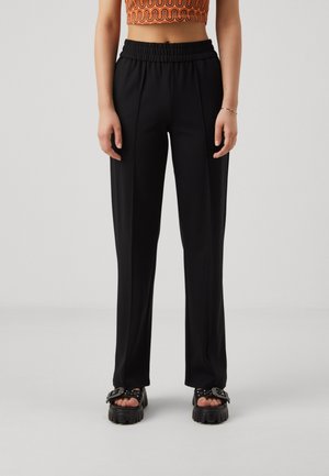 Femme portant un pantalon noir taille haute, coupe droite, avec une ceinture élastique et des sandales noires épaisses avec de grandes boucles.