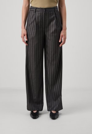 Kvinde iført sort, vidde-bukser med højtaljede og hvide, lodrette nålestriber samt sorte spidse sko, stående mod en lys grå baggrund.