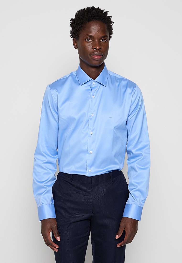 Hellblaues langärmliges Hemd aus glattem Stoff, mit spitzem Kragen und weißen Knöpfen, getragen zu dunklen Hosen.