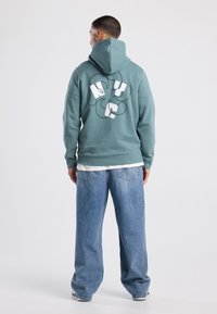 Mintgroene hoodie met een witte grafische print op de achterkant, gecombineerd met lichtblauwe denim jeans in ontspannen pasvorm.