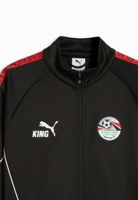 Veste de sport noire zippée avec des bandes d'épaule rouges à motifs, logo Puma blanc et texte « KING » sur la poitrine gauche, et emblème de la Fédération égyptienne de football à droite.
