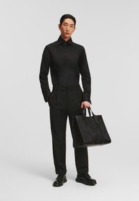 Čierna košeľa na mieru s hladkou textúrou v kombinácii s čiernymi nohavicami. K outfitu patrí aj textúrovaná čierna kabelka s dvoma úchytmi.