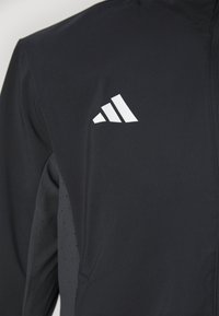 Veste de sport noire avec logo blanc à trois bandes sur la poitrine et panneaux latéraux perforés, équipée d'une fermeture éclair à l'avant.