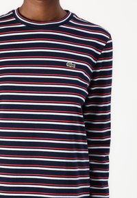 Jūrinių ilgesnių rankovių marškiniai su horizontaliomis raudonomis ir baltomis juostelėmis, priekyje esantis mažas žalias Lacoste logotipas. Pagaminta iš medvilnės, su apvaliu kaklu.