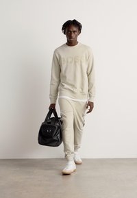 Sudadera de color beige claro con el logo 'BOSS', joggers a juego, camiseta blanca debajo y zapatillas blancas. Lleva un bolso de deporte negro.