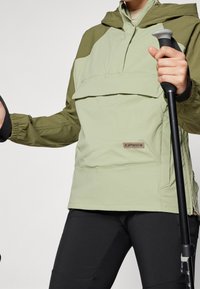 Chaqueta con capucha de color verde claro con acentos en verde más oscuro, bolsillos laterales con cremallera y puños elásticos, combinada con pantalones negros y un bastón de senderismo.