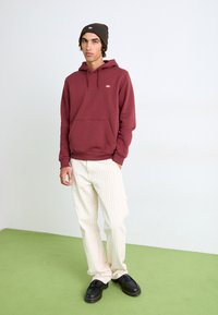 Bordeaux hættebluse med en frontlomme og logo, parret med crème-stribede bukser og sorte sko. Inkluderer en mørkegrøn strikket hat.