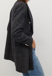 Dlouhý černý a šedý kostkovaný blazer s uvolněným střihem, zdobení zlatými knoflíky a dvěma předními kapsami, v kombinaci s modrými džíny.