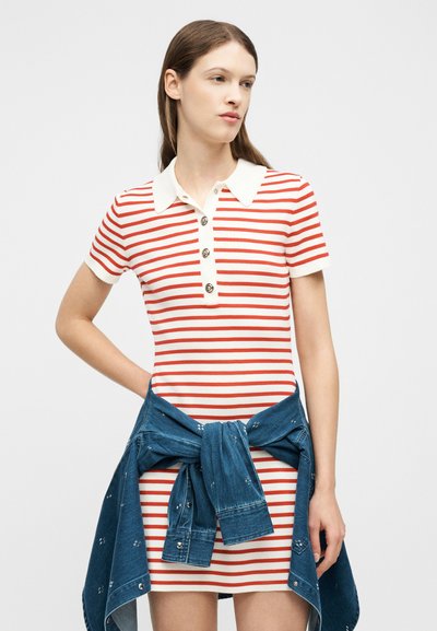 Γυναίκα που φοράει κόκκινο και άσπρο ριγέ φόρεμα πόλο με λευκό γιακά, τζιν μπουφάν δεμένο γύρω από τη μέση, στέκεται μπροστά σε απλό φόντο.