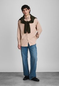 Giovane uomo in piedi con le mani dietro la schiena, indossa una camicia beige con bottoni, jeans blu, scarpe nere e un maglione verde scuro annodato sulle spalle.