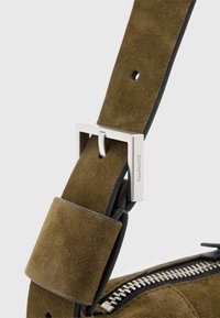 Correa de bolsa de ante verde con bordes negros, que presenta una hebilla de metal plateado y un logotipo. La correa tiene una textura suave y longitud ajustable.