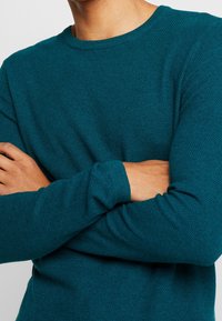 Teal texturerad stickad tröja med rundad halsringning och långa ärmar, med ribbade muddar och en figurnära silhuett.