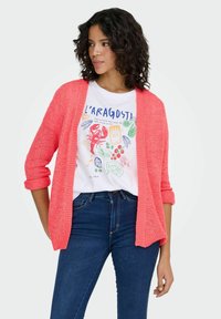 Bright coral knit cardigan with rolled sleeves, worn over a white graphic t-shirt featuring colorful food illustrations and dark blue jeans.