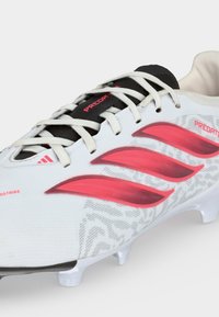 Λευκό ποδοσφαιρικό παπούτσι με ροζ ρίγες Adidas, μαύρη γλώσσα με την ένδειξη "Predator" και λευκά κορδόνια με διαμορφωμένες προστασίες στη σόλα.