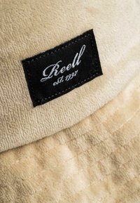 Beige strukturiertes Gewebe mit einem schwarz eingestickten Etikett, auf dem in weißer Kursivschrift "Reell est. 1997" steht.