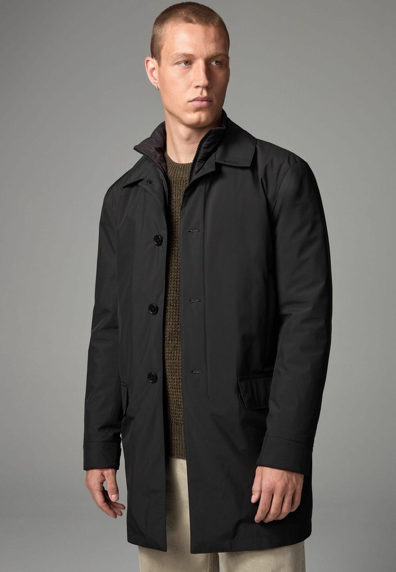 Abrigo negro impermeable con cierre de botón, cuello con muesca, bolsillos laterales y una textura suave, superpuesto a un suéter de punto verde oscuro.