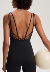 Čierny jednodielny plavky s prekríženými ramenami a hladkým povrchom, držiacím texturovanú krémovú guľu.