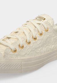 Ανοιχτόχρωνο (ελαφρώς υπόλευκο) παπούτσι Converse με υφασμένο λουλουδένιο σχέδιο, κρεμ κορδόνια, χρυσά περπάκια και χρυσό λογότυπο Converse Star στη γλώσσα.