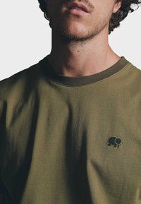 Camiseta de algodón verde oliva con escote redondo, que presenta un pequeño bordado de un elefante negro en el área del pecho izquierdo. Textura suave.