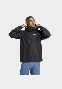 Giacca impermeabile nera con zip frontale e cappuccio regolabile, realizzata in materiale leggero, dotata di due tasche laterali e dettagli del logo.