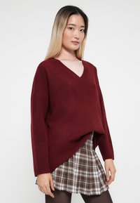 Bordeaux oversized sweater met een diepe V-hals en lange mouwen, gecombineerd met een bruine geruite mini-jurk. Het materiaal lijkt zacht en textuurrijk.