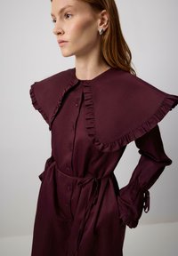 Burgundy klänning med volangkrage och manschetter, knäppning framtill och knyt i midjan. Tillverkad av slätt tyg, har en strukturerad design och eleganta detaljer.