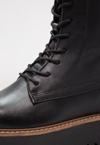 Bota de tobillo de cuero negro con cordones planos, textura suave, costuras de acento y una suela de plataforma de color marrón claro contrastante.