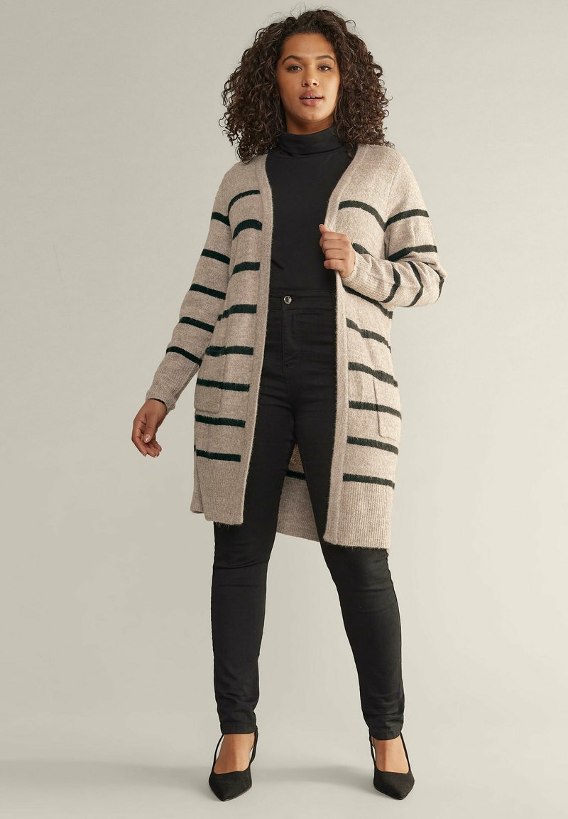 Lång beige cardigan med mörkgröna ränder, öppen front och ribbade manschetter, lager-på-lager över en svart turtleneck och svarta tajta byxor.