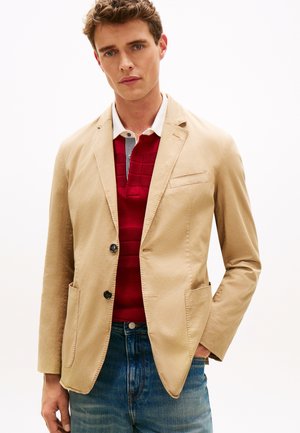 Man met beige colbert over een rood poloshirt en blauwe spijkerbroek, staand met één hand in zijn zak tegen een effen achtergrond.