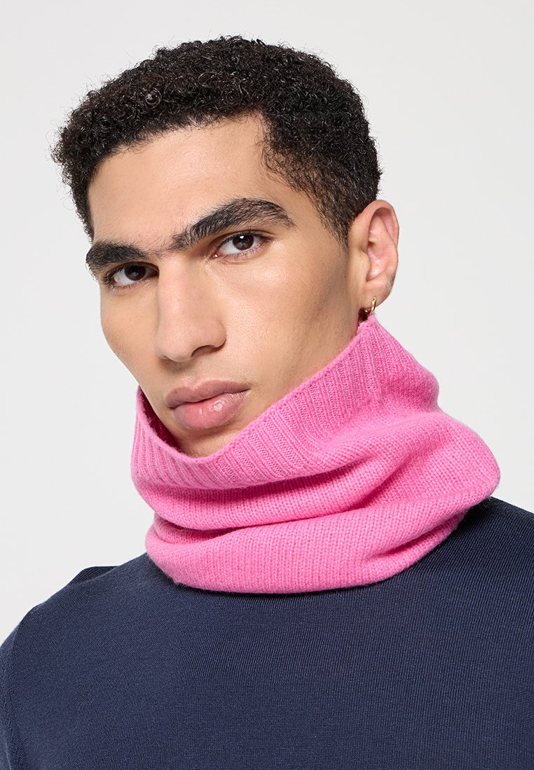 Różowy szydełkowany szalik o prążkowanej strukturze, noszony w złożonym stylu wokół szyi. Model ma na sobie granatową koszulę.