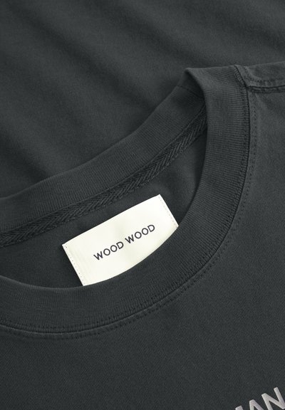 Tumši pelēks kokvilnas T-krekls ar apaļu kakla izgriezumu, redzamām šuvēm un uzlīmi ar uzrakstu "WOOD WOOD" netālu no apkakles.