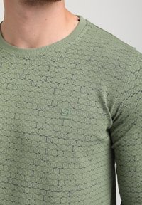 Suéter verde de punto con textura geométrica, cuello circular y un pequeño logo en el pecho. La tela presenta un patrón sutil con acentos oscuros.