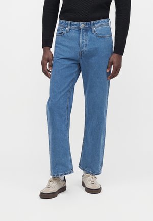 Homme portant un jean droit bleu clair, une chemise noire texturée à manches longues et des baskets beiges à semelles foncées sur un fond blanc.