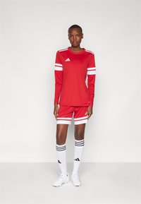 Maglietta sportiva a maniche lunghe rossa e pantaloni corti con strisce bianche, abbinati a calze bianche con righe nere e scarpe da calcio bianche.
